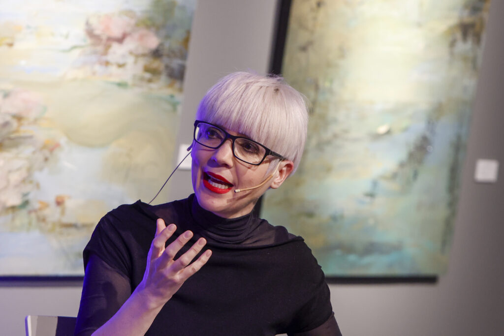 Woman with short platinum blonde hair and glasses speaks, gesturing with her hands in front of abstract wall art, wearing a black top with a headset mic.